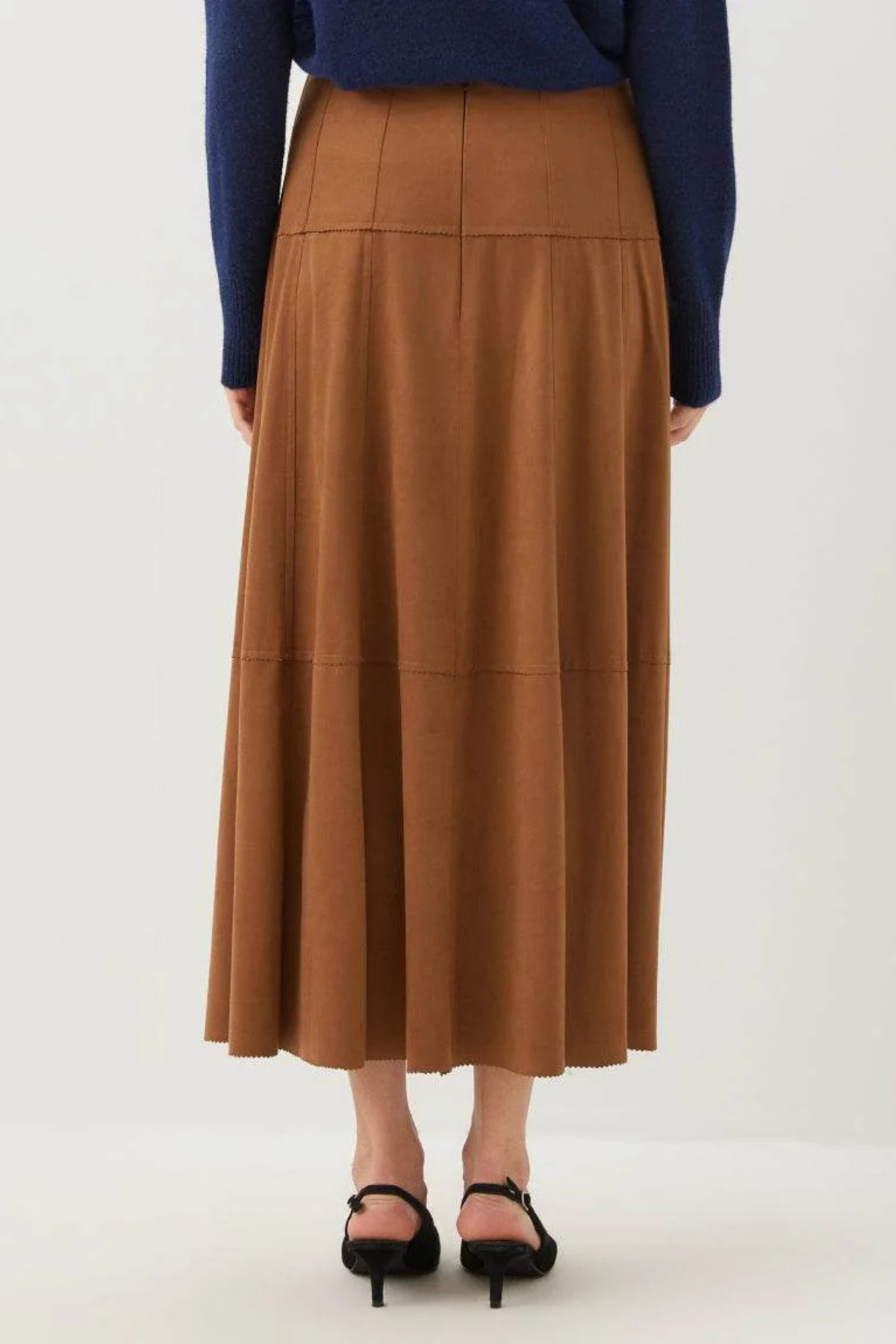 A model is shown from the back wearing a brown pleated midi skirt, a navy blue long-sleeve top, and black heeled sandals against a plain white studio background.