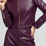 A woman wearing a shiny dark purple faux leather jacket and matching shorts with gold-tone buttons and zipper details, shown against a light gray studio background.