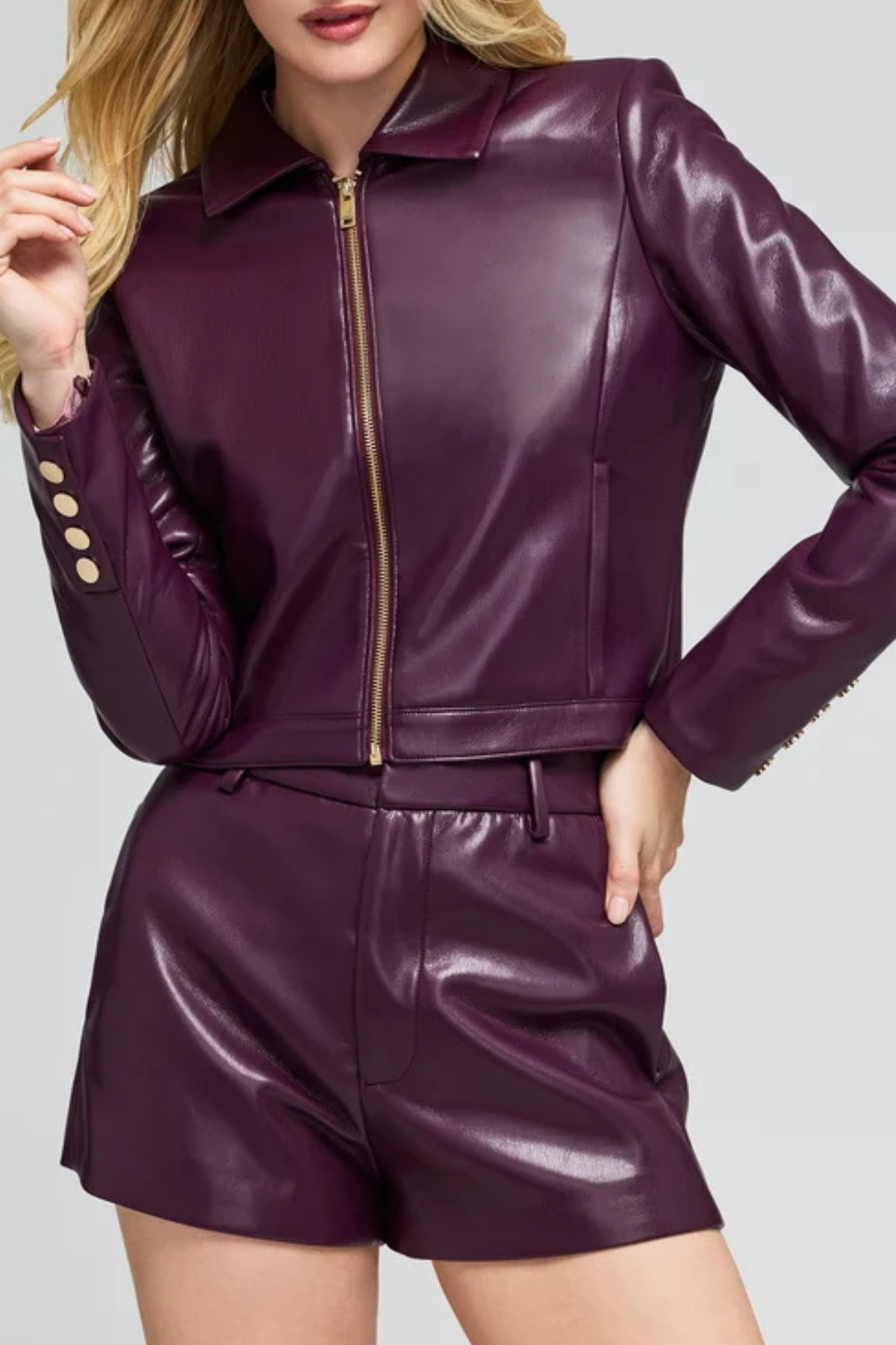 A woman wearing a shiny dark purple faux leather jacket and matching shorts with gold-tone buttons and zipper details, shown against a light gray studio background.