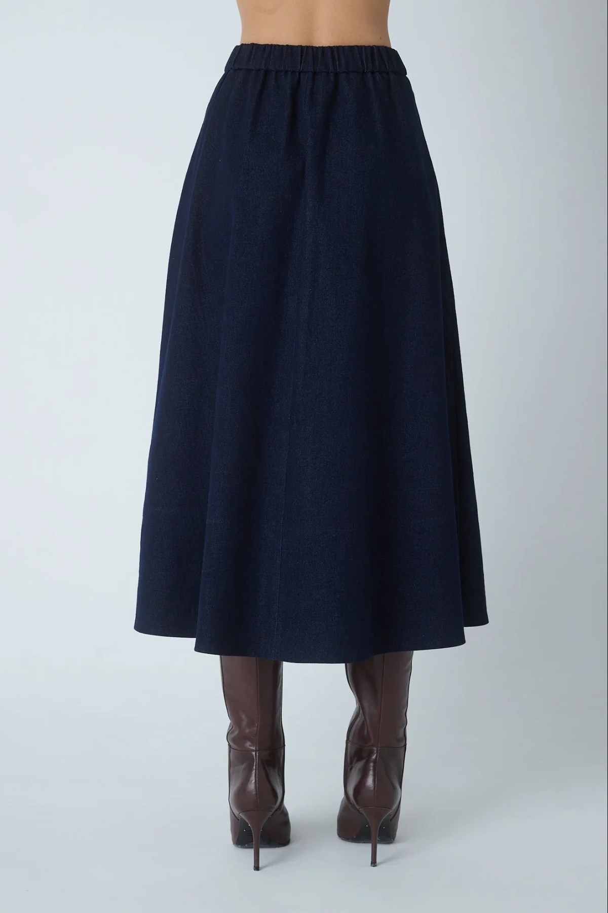 A dark navy blue A-line midi skirt with an elastic waistband, shown on a model from the back wearing brown high-heeled boots against a light gray studio background.