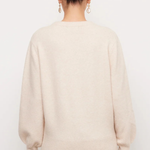 A woman viewed from the back wearing a cream-colored knit sweater and dark pants against a plain white background.