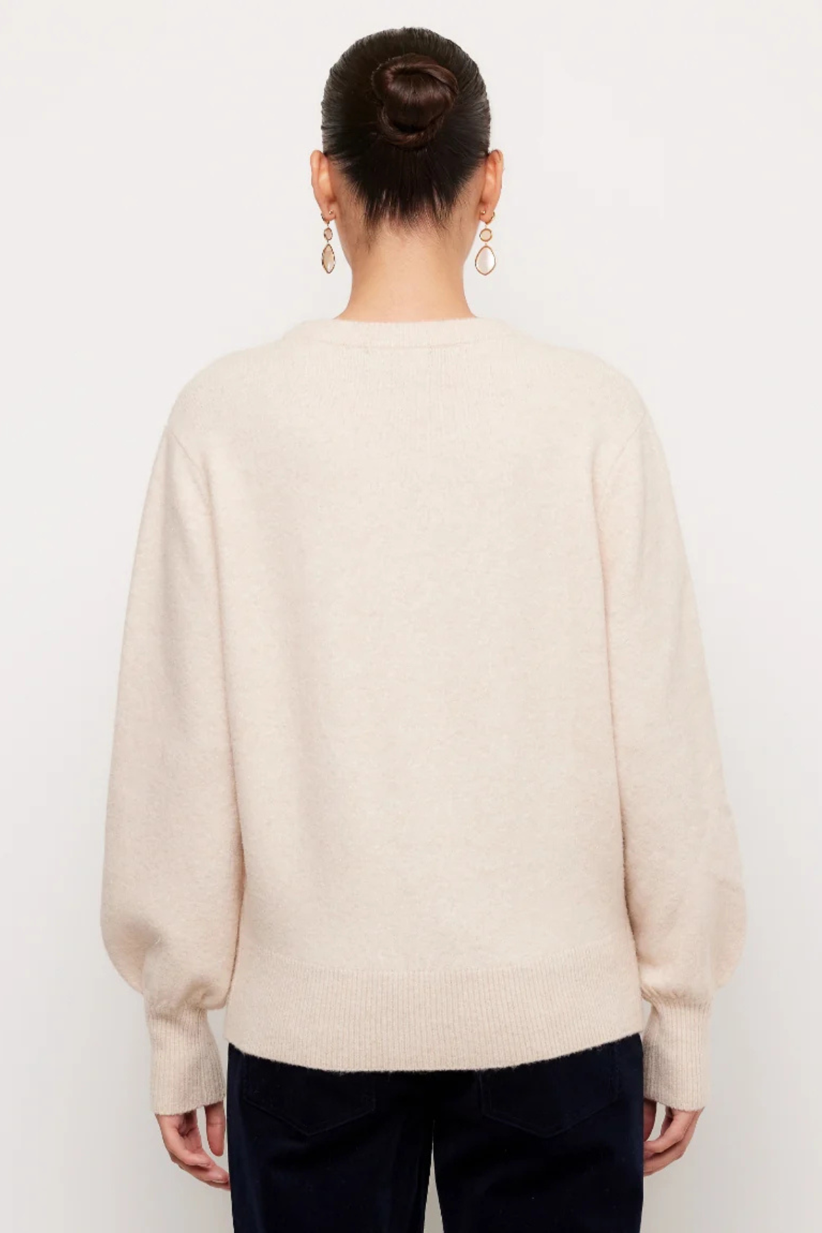 A woman viewed from the back wearing a cream-colored knit sweater and dark pants against a plain white background.
