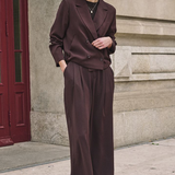 A woman stands on a city sidewalk wearing a tailored two-piece suit in a deep chocolate brown satin-like fabric, featuring a double-breasted blazer and matching wide-leg trousers.