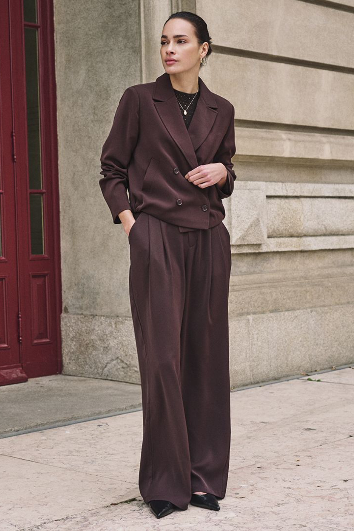 A woman stands on a city sidewalk wearing a tailored two-piece suit in a deep chocolate brown satin-like fabric, featuring a double-breasted blazer and matching wide-leg trousers.