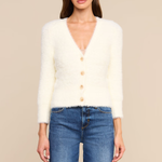 A woman modeling a fuzzy off-white cardigan sweater with a V-neck and three front buttons, paired with blue jeans, against a solid beige background.