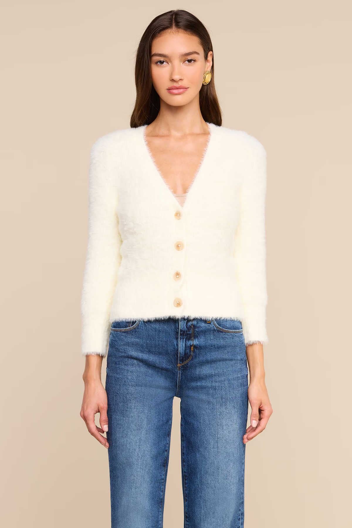 A woman modeling a fuzzy off-white cardigan sweater with a V-neck and three front buttons, paired with blue jeans, against a solid beige background.