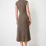 A woman stands with her back to the camera wearing a sleeveless, ribbed knit midi dress in a taupe-brown color, paired with black pointed-toe flats, against a plain white wall and light concrete floor.