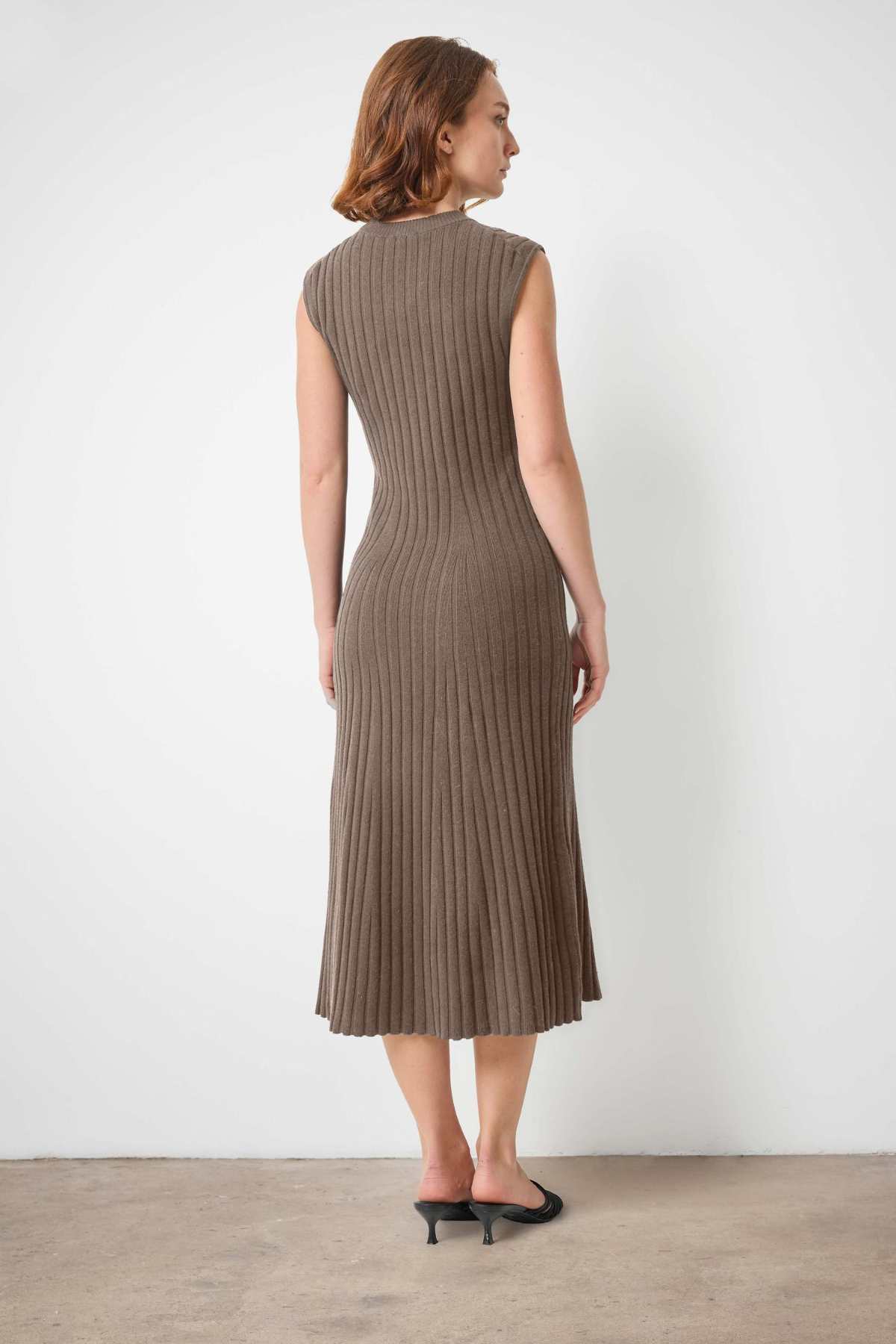 A woman stands with her back to the camera wearing a sleeveless, ribbed knit midi dress in a taupe-brown color, paired with black pointed-toe flats, against a plain white wall and light concrete floor.