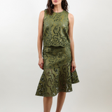 A woman modeling a sleeveless olive green midi dress with a textured, patterned surface and a ruffled hem, paired with brown slide sandals, against a plain light gray studio background.