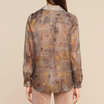 A woman viewed from the back wearing a long-sleeve button-up shirt with an abstract geometric print in brown, beige, and gold tones, paired with light beige pants, against a solid beige background.