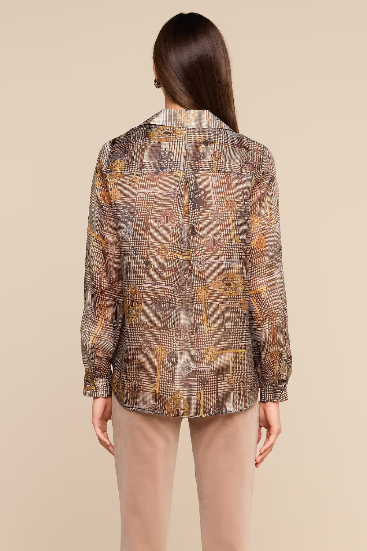 A woman viewed from the back wearing a long-sleeve button-up shirt with an abstract geometric print in brown, beige, and gold tones, paired with light beige pants, against a solid beige background.