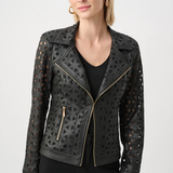 A woman wearing a black faux-leather moto jacket with geometric laser-cutout patterns and gold-tone zippers, paired with a black top and black pants, against a light gray studio background.