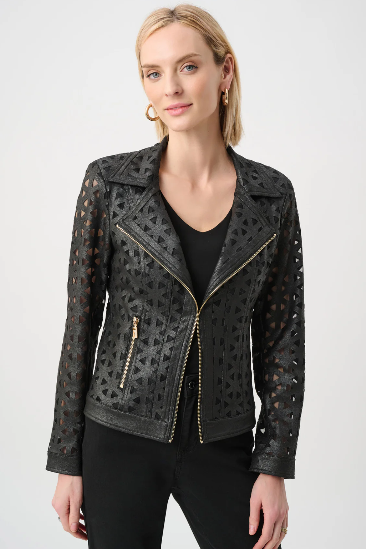 A woman wearing a black faux-leather moto jacket with geometric laser-cutout patterns and gold-tone zippers, paired with a black top and black pants, against a light gray studio background.