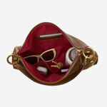An open brown leather handbag with a deep red interior, shown against a light gray background. Inside, beige sunglasses, a small white bottle, and keys are arranged in the compartment.