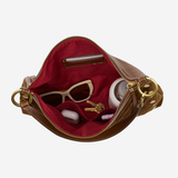 An open brown leather handbag with a deep red interior, shown against a light gray background. Inside, beige sunglasses, a small white bottle, and keys are arranged in the compartment.