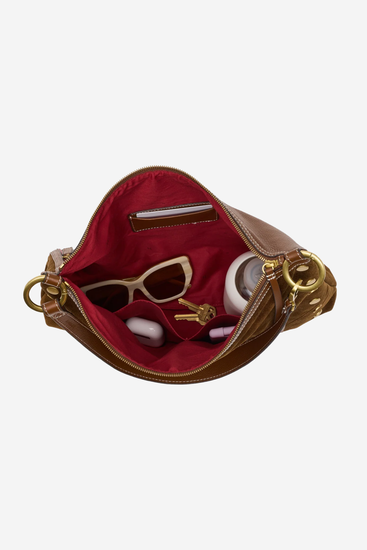 An open brown leather handbag with a deep red interior, shown against a light gray background. Inside, beige sunglasses, a small white bottle, and keys are arranged in the compartment.
