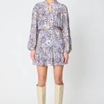 A model stands against a light gray studio background wearing a long-sleeve mini dress with a blue, orange, and white paisley pattern, paired with beige knee-high boots.
