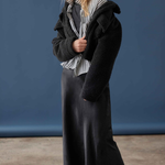A woman models a black fuzzy cropped jacket over a black and white striped shirt, a long black skirt, and dark purple tights against a blue studio backdrop.