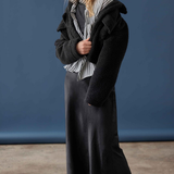 A woman models a black fuzzy cropped jacket over a black and white striped shirt, a long black skirt, and dark purple tights against a blue studio backdrop.