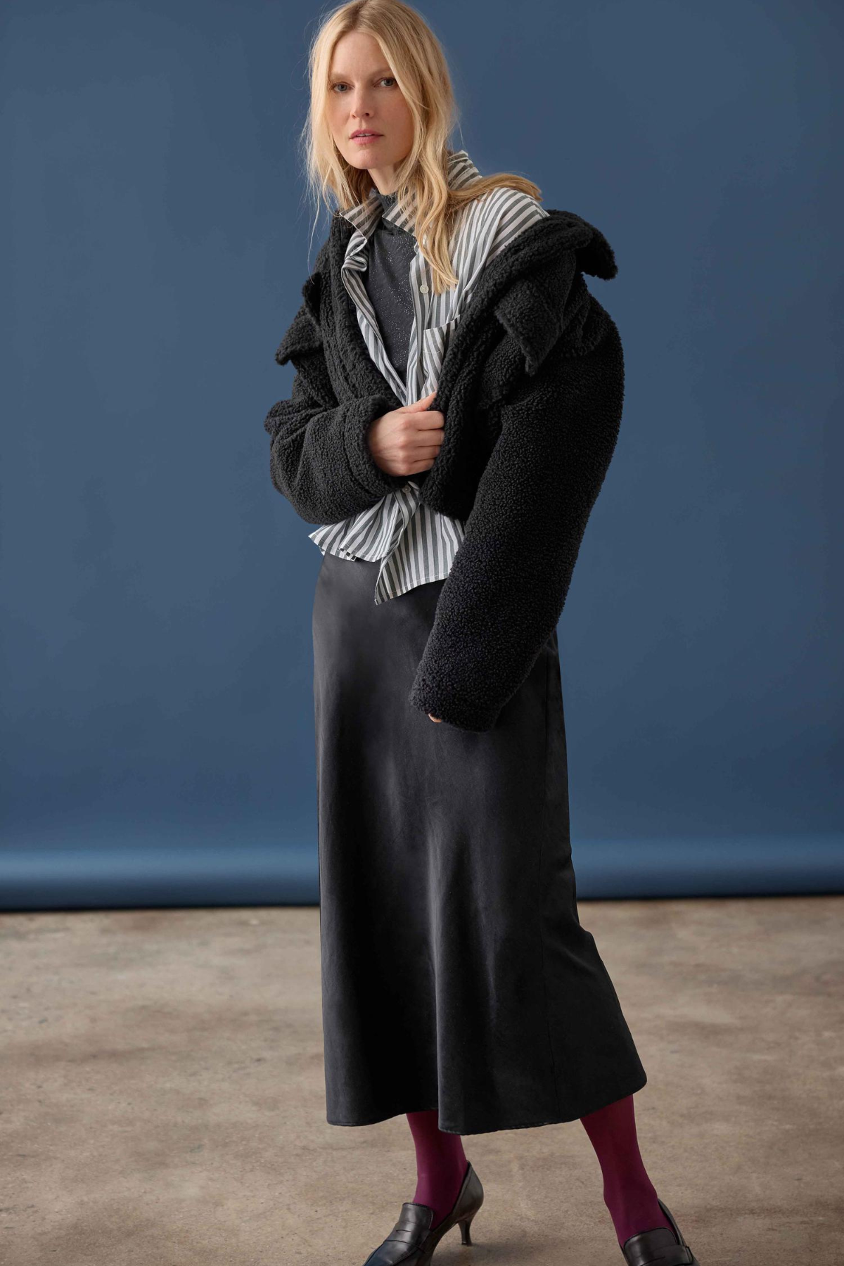 A woman models a black fuzzy cropped jacket over a black and white striped shirt, a long black skirt, and dark purple tights against a blue studio backdrop.