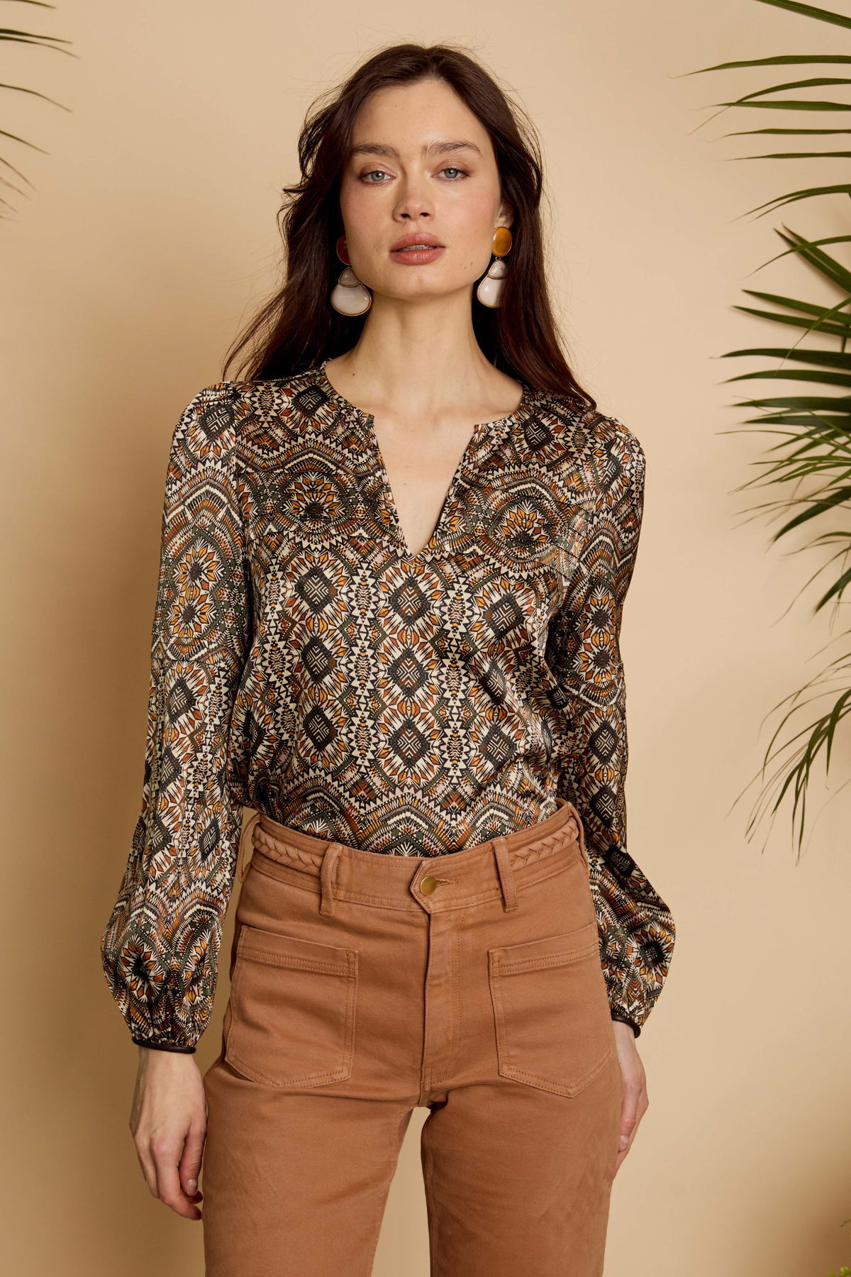 Woman wearing a patterned blouse and brown pants against a beige background
