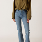 A model wearing an olive green long-sleeve button-up shirt and light-wash blue cropped jeans with black leather shoes, standing against a plain off-white studio background.