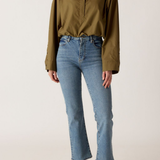 A model wearing an olive green long-sleeve button-up shirt and light-wash blue cropped jeans with black leather shoes, standing against a plain off-white studio background.