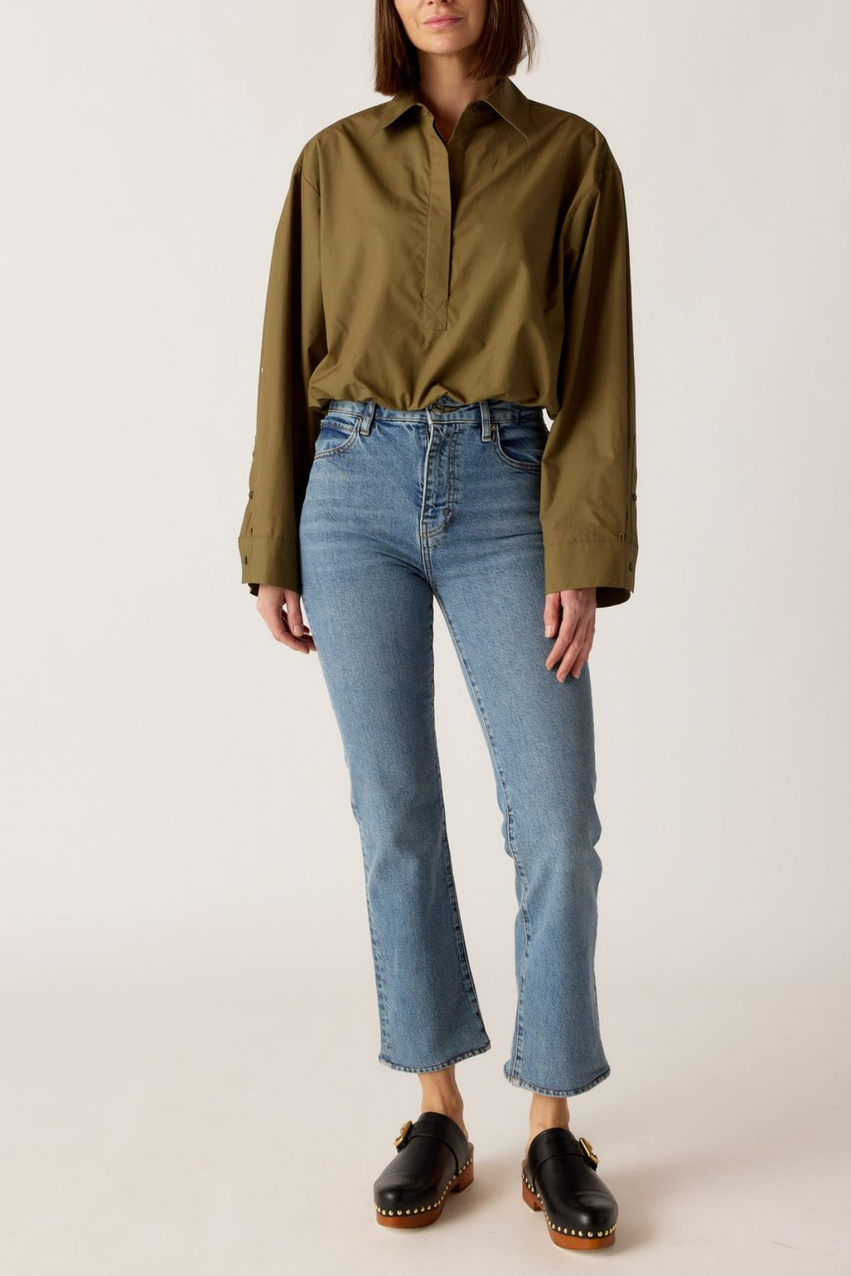 A model wearing an olive green long-sleeve button-up shirt and light-wash blue cropped jeans with black leather shoes, standing against a plain off-white studio background.