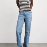 A model wearing a black and white striped sleeveless top, light blue straight-leg jeans, and tan sneakers with green laces, standing against a plain light gray studio background.