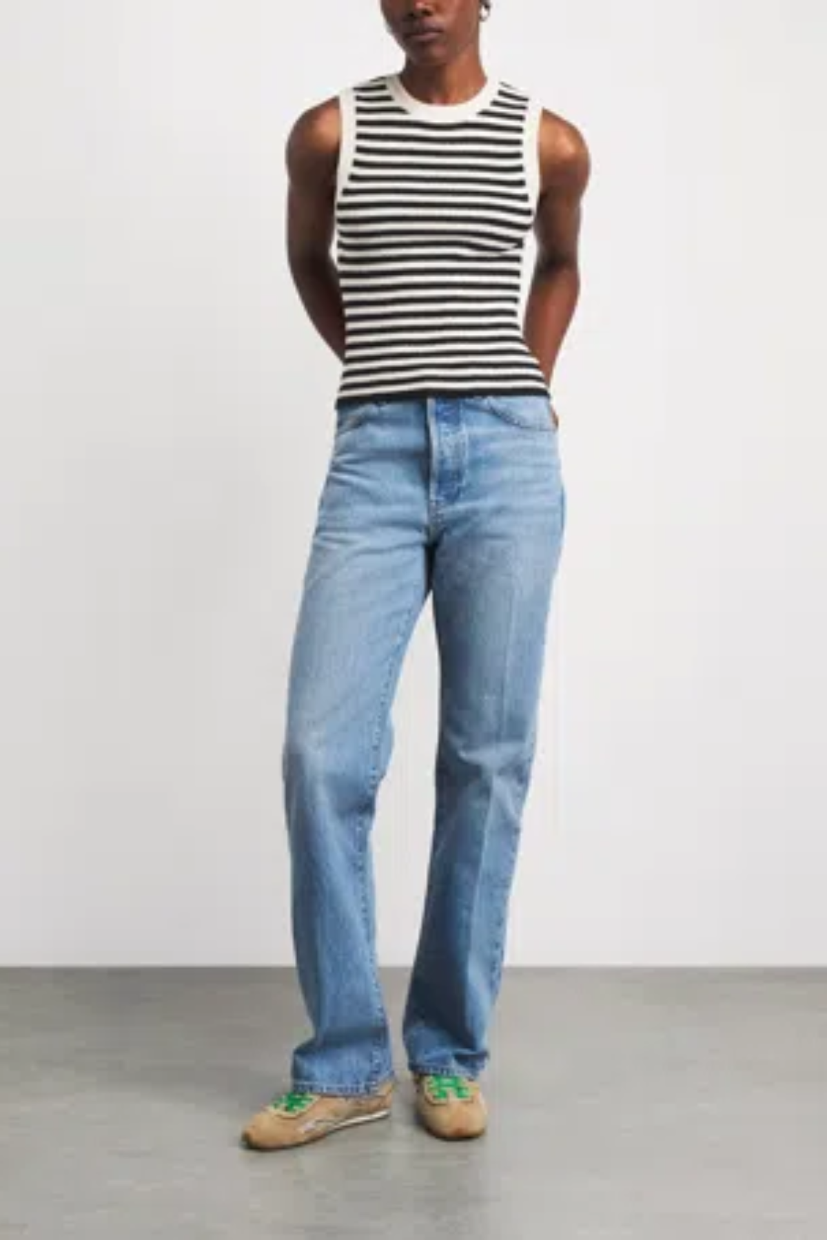 A model wearing a black and white striped sleeveless top, light blue straight-leg jeans, and tan sneakers with green laces, standing against a plain light gray studio background.