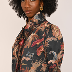 A woman with a large black afro hairstyle wearing a short-sleeved button-up shirt with a dark tropical floral print in beige, red, and blue, paired with gold hoop earrings, against a solid light beige background.