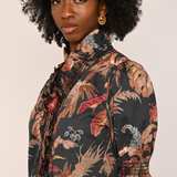 A woman with a large black afro hairstyle wearing a short-sleeved button-up shirt with a dark tropical floral print in beige, red, and blue, paired with gold hoop earrings, against a solid light beige background.