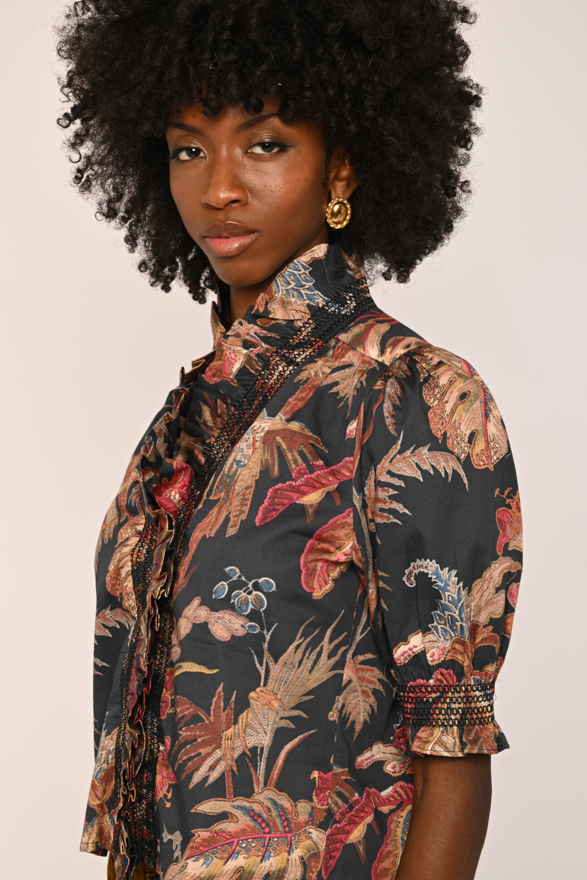 A woman with a large black afro hairstyle wearing a short-sleeved button-up shirt with a dark tropical floral print in beige, red, and blue, paired with gold hoop earrings, against a solid light beige background.