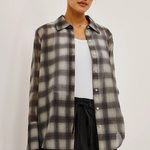 A woman wearing a semi-sheer long-sleeve button-up plaid shirt in gray, black, and white, layered over a white tank top and black pants, set against a plain light background.