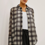 A woman wearing a semi-sheer long-sleeve button-up plaid shirt in gray, black, and white, layered over a white tank top and black pants, set against a plain light background.