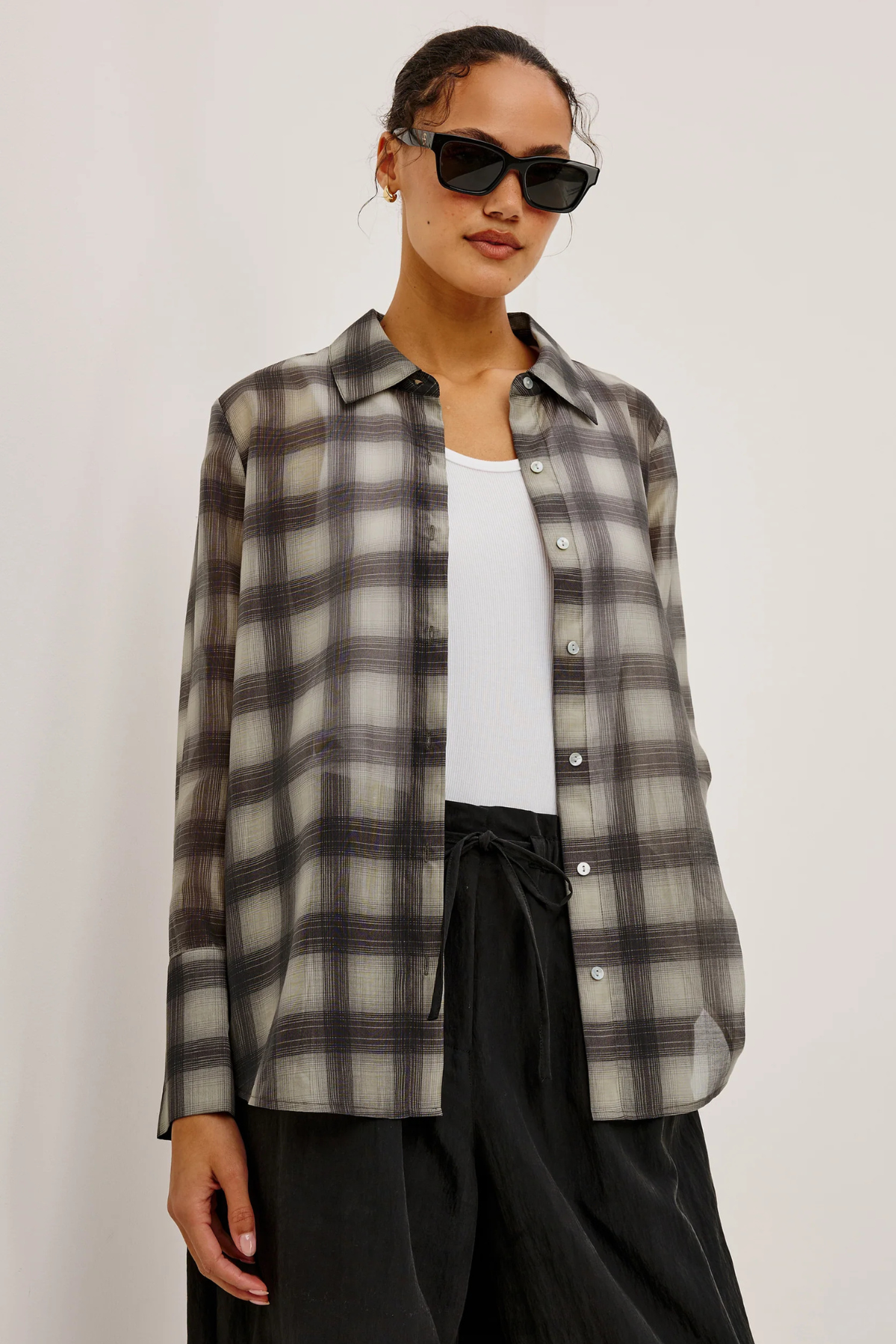 A woman wearing a semi-sheer long-sleeve button-up plaid shirt in gray, black, and white, layered over a white tank top and black pants, set against a plain light background.
