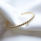 A gold-toned twisted rope cuff bracelet resting on draped white fabric.