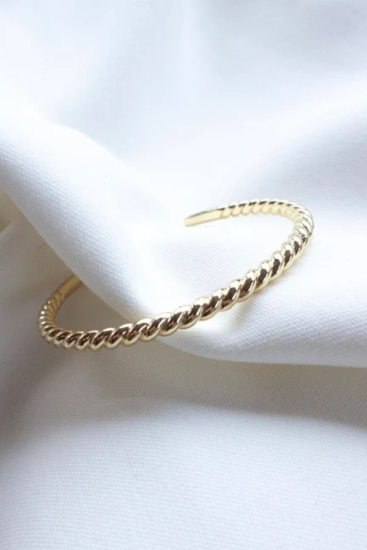 A gold-toned twisted rope cuff bracelet resting on draped white fabric.