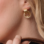 A close-up of a model's ear wearing a gold hoop earring with a textured, beaded outer ring and a smaller silver stud earring. The model has blonde hair and is wearing gold rings on her fingers.