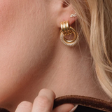 A close-up of a model's ear wearing a gold hoop earring with a textured, beaded outer ring and a smaller silver stud earring. The model has blonde hair and is wearing gold rings on her fingers.