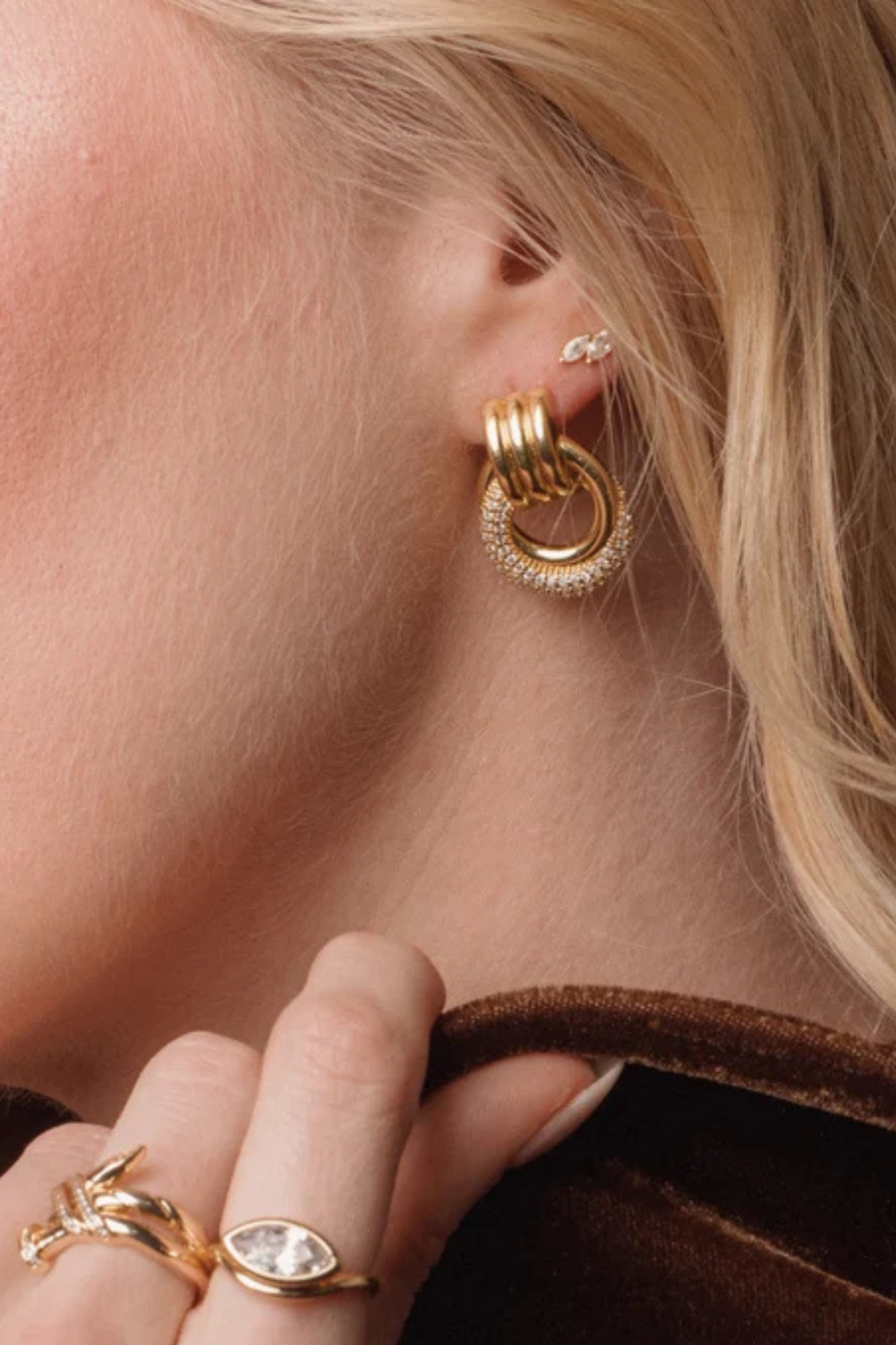 A close-up of a model's ear wearing a gold hoop earring with a textured, beaded outer ring and a smaller silver stud earring. The model has blonde hair and is wearing gold rings on her fingers.
