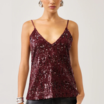 A woman modeling a burgundy sequined camisole top with thin spaghetti straps and a V-neckline, paired with black pants, against a light gray studio background.