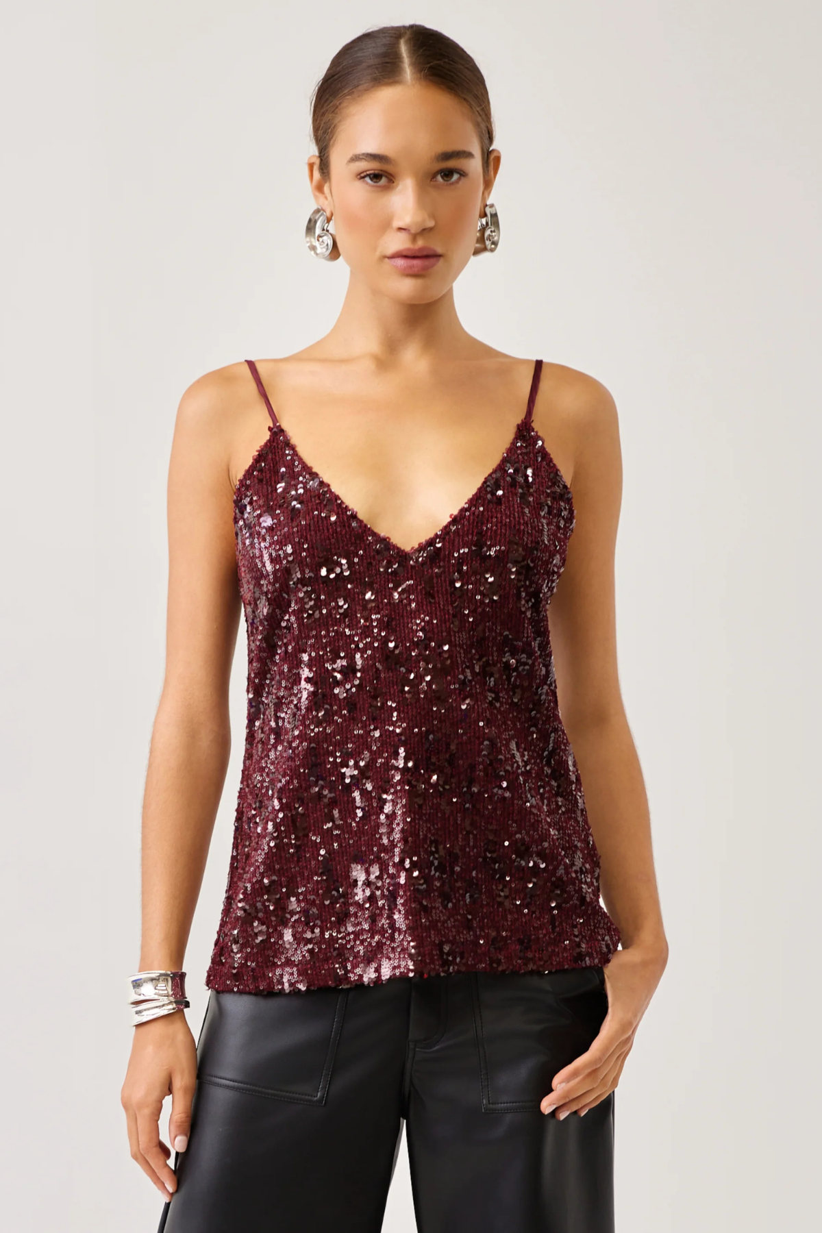 A woman modeling a burgundy sequined camisole top with thin spaghetti straps and a V-neckline, paired with black pants, against a light gray studio background.