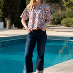A woman stands by a swimming pool wearing a short-sleeve button-up blouse with a multicolor floral and geometric print, dark cuffed jeans, and navy blue flats.