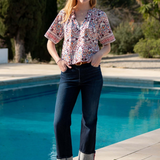 A woman stands by a swimming pool wearing a short-sleeve button-up blouse with a multicolor floral and geometric print, dark cuffed jeans, and navy blue flats.