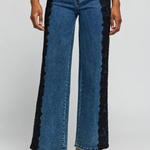 A model wears blue wide-leg jeans with black lace panels running down the outer sides, styled with a navy top and black heels against a light gray studio background.