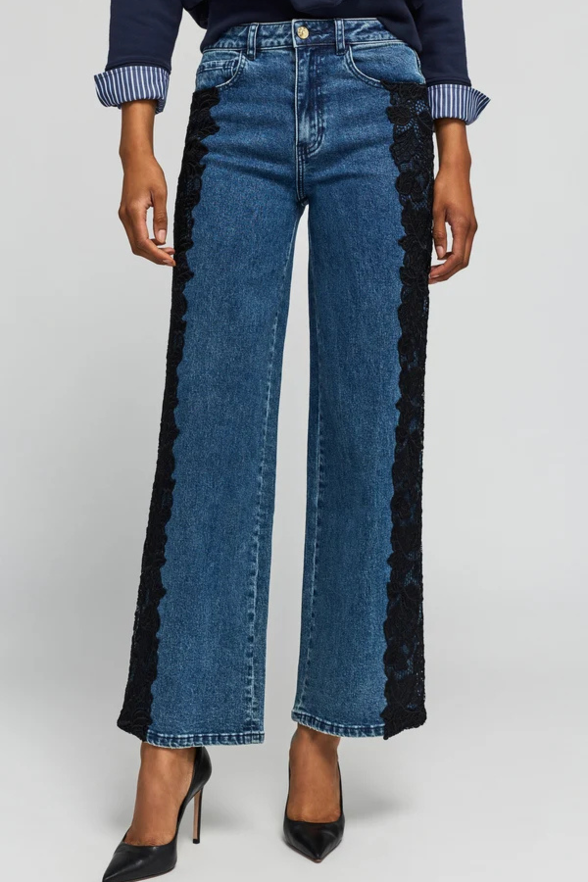 A model wears blue wide-leg jeans with black lace panels running down the outer sides, styled with a navy top and black heels against a light gray studio background.
