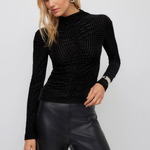 A woman modeling a black textured long-sleeve mock neck top paired with black faux leather pants, against a light gray studio background.