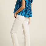 back of woman in blue floral blouse and white pants on tan background 