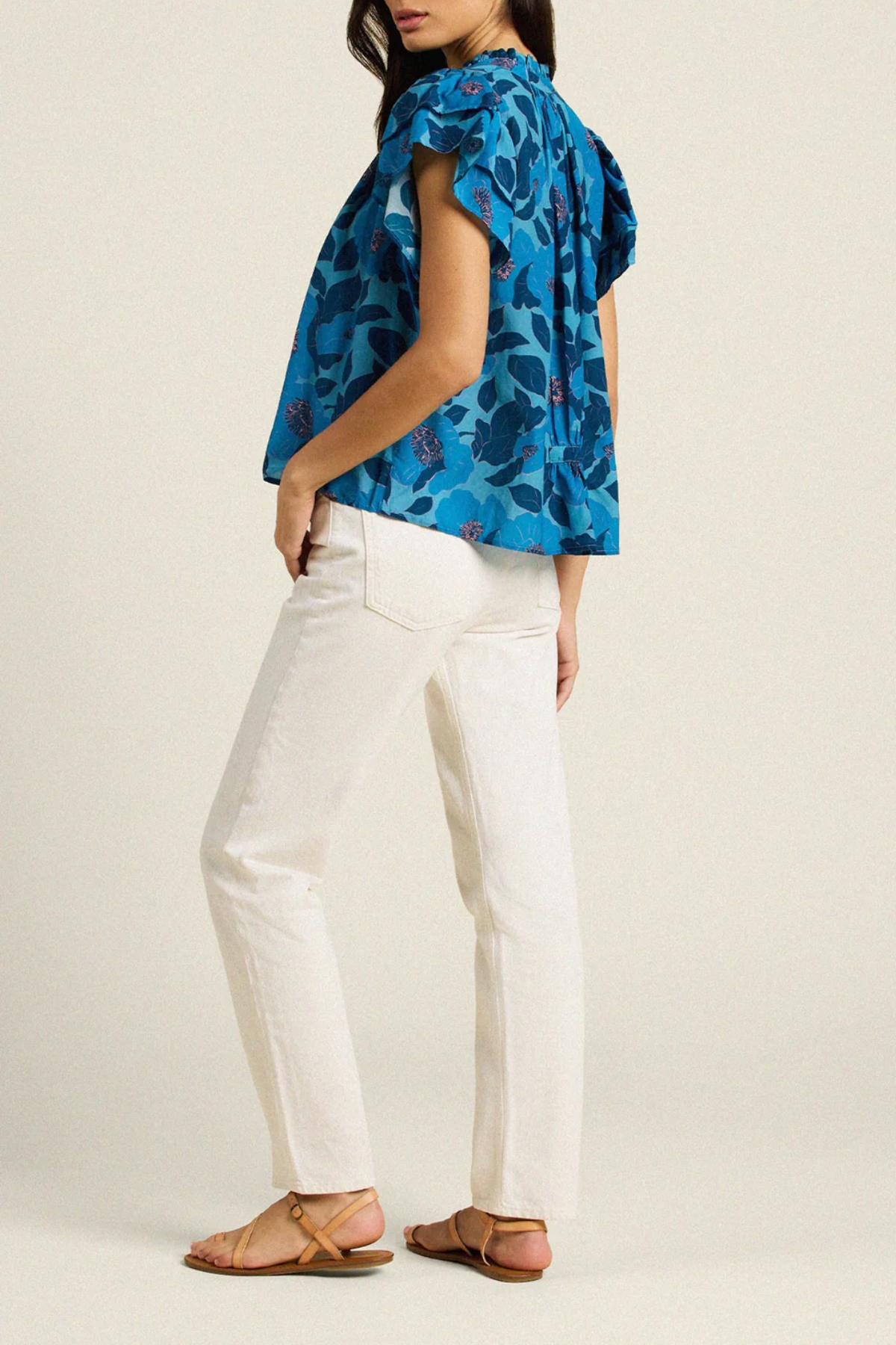 back of woman in blue floral blouse and white pants on tan background 